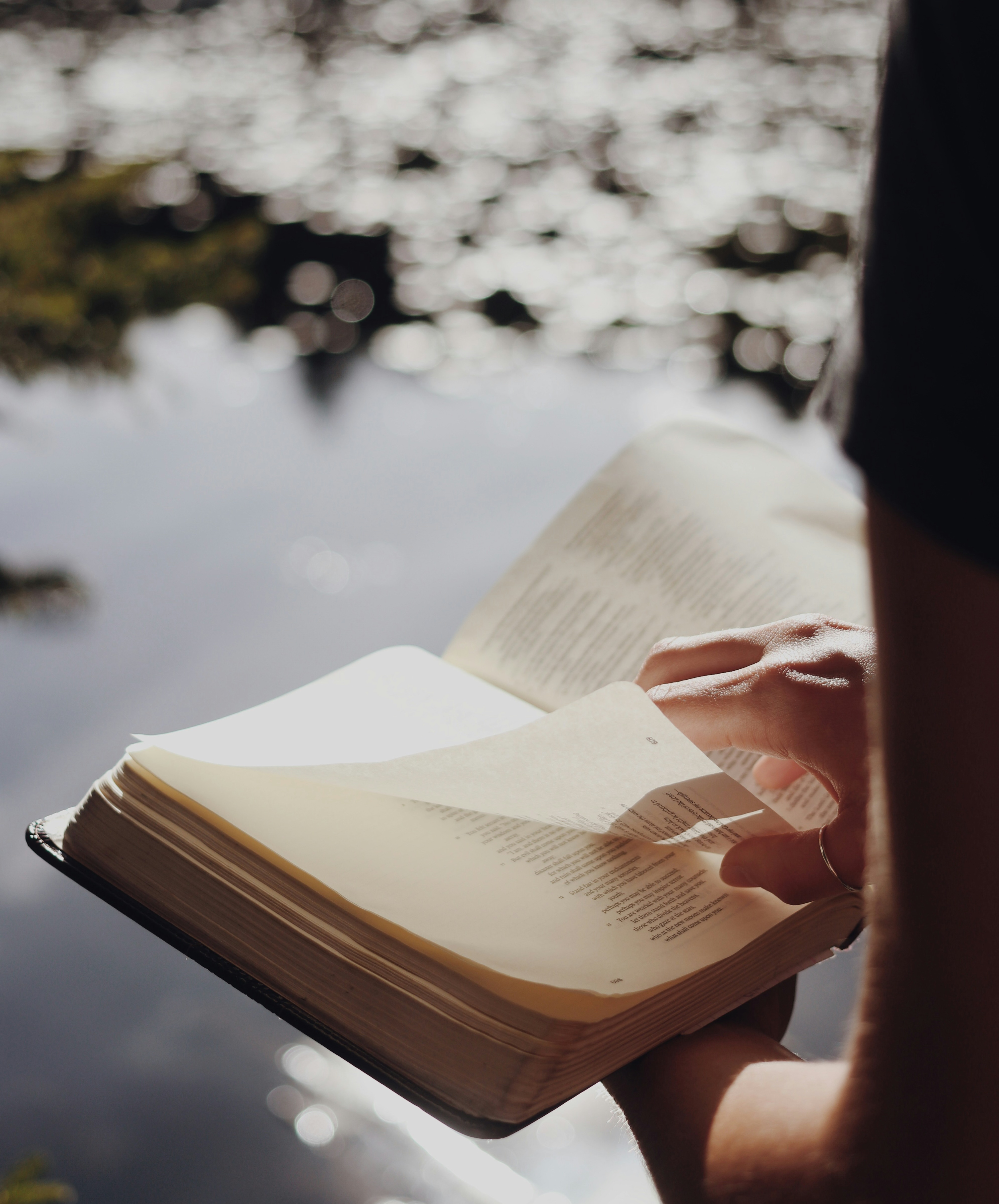 A pair of hands hold a Bible against a backdrop of a body of water on a sunny day.