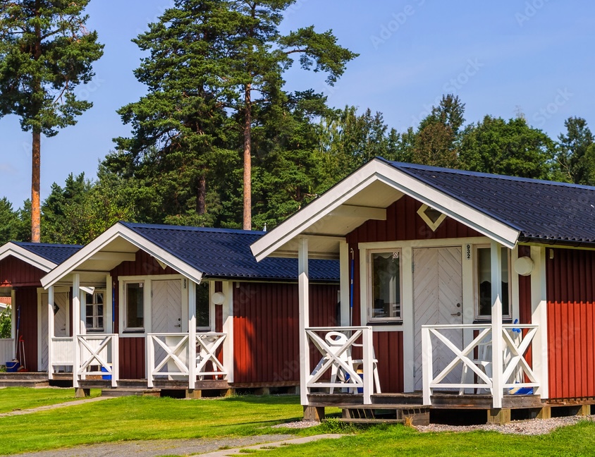 three summer camp cabins on a manicured lawn against the backdrop of a pine forest on a sunny day