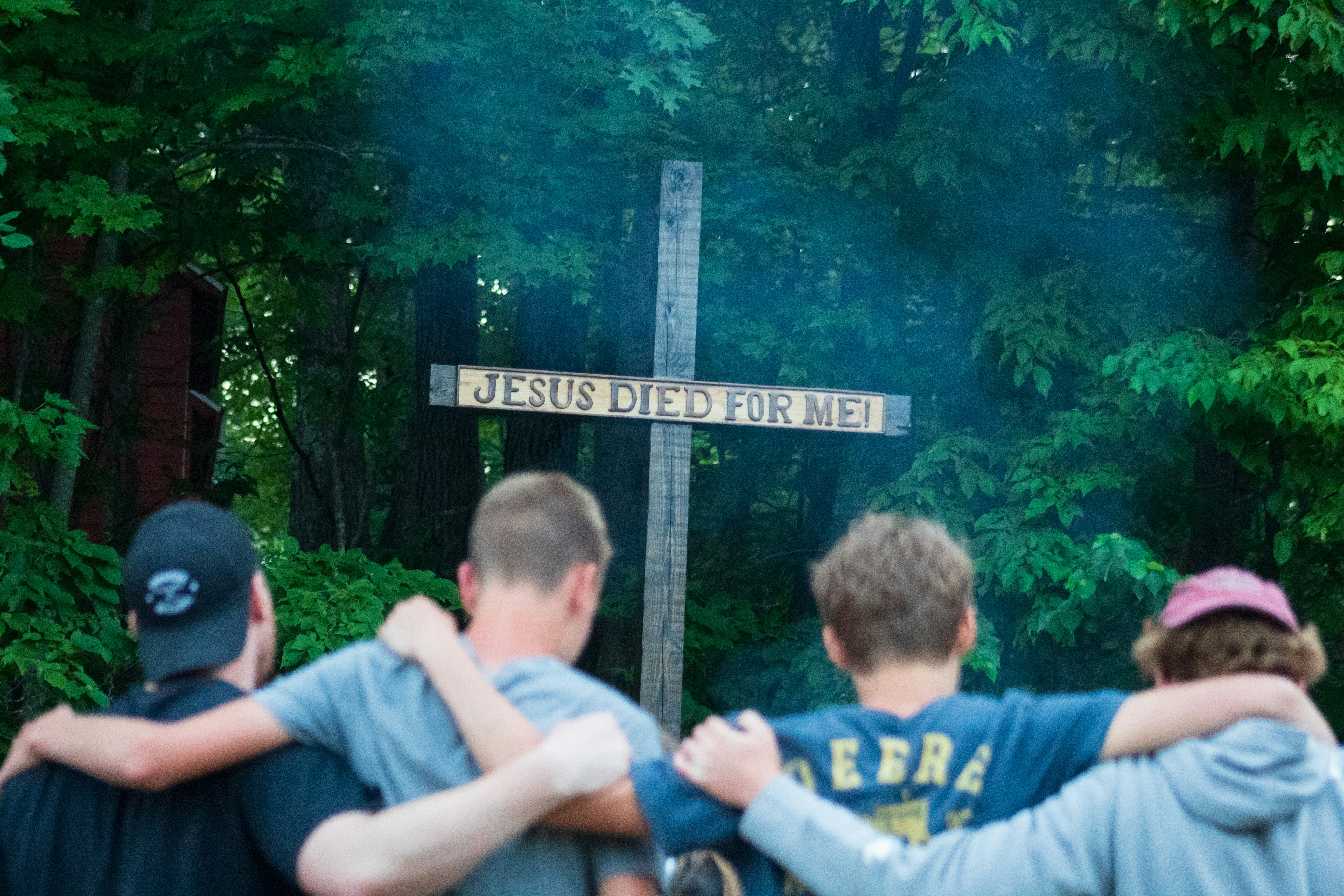 Four teenage boys stand with their backs to the camera, their arms around each others shoulders. The are looking at a large wooden cross carved with the words `JESUS DIED FOR ME!`